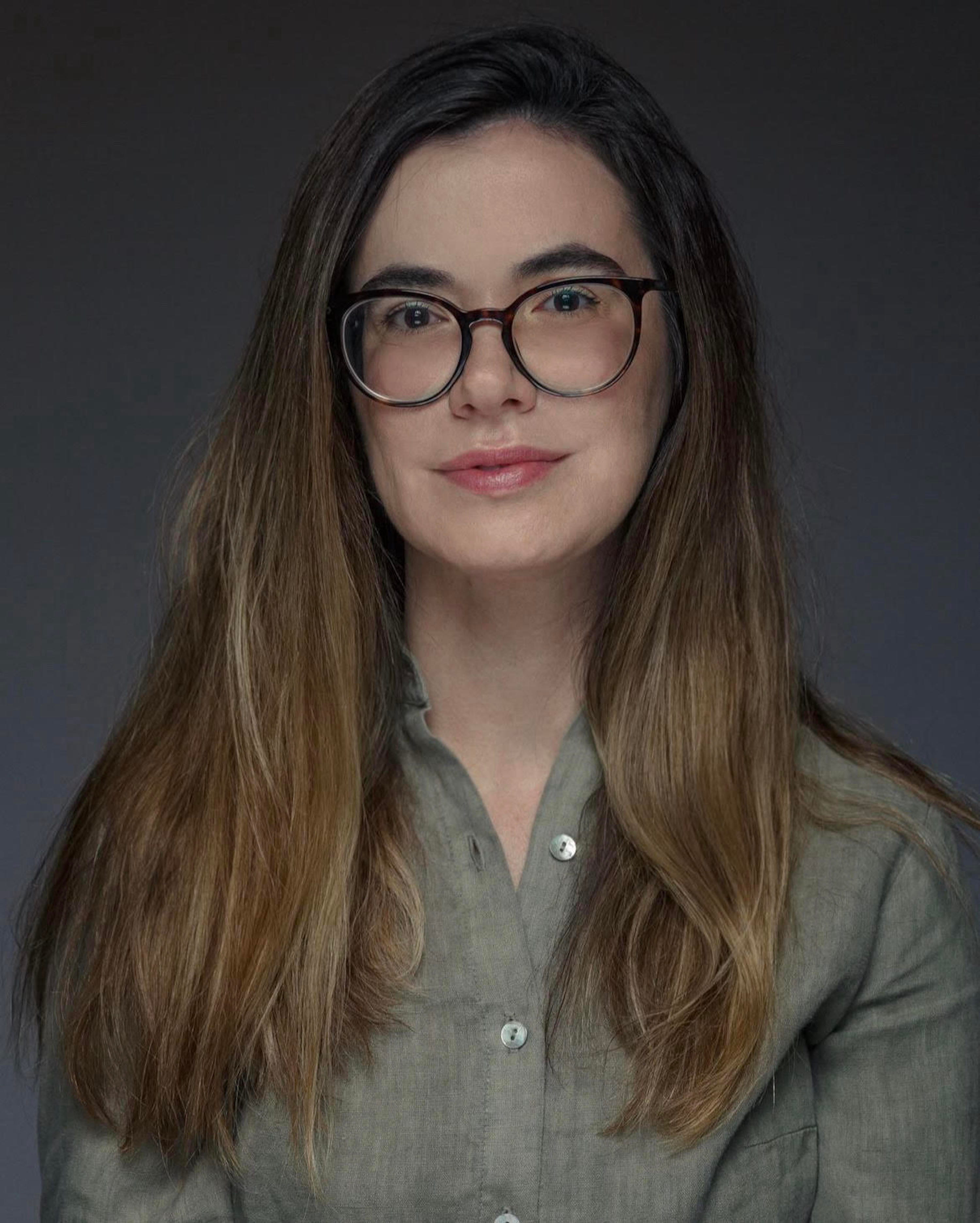 Fotografia de Fabiane Secches. Uma mulher branca, de &oacute;culos, com o cabelo castanho com as pontas loiras. Ela veste uma camisa social verde musgo. O fundo &eacute; um gradiente preto e cinza.